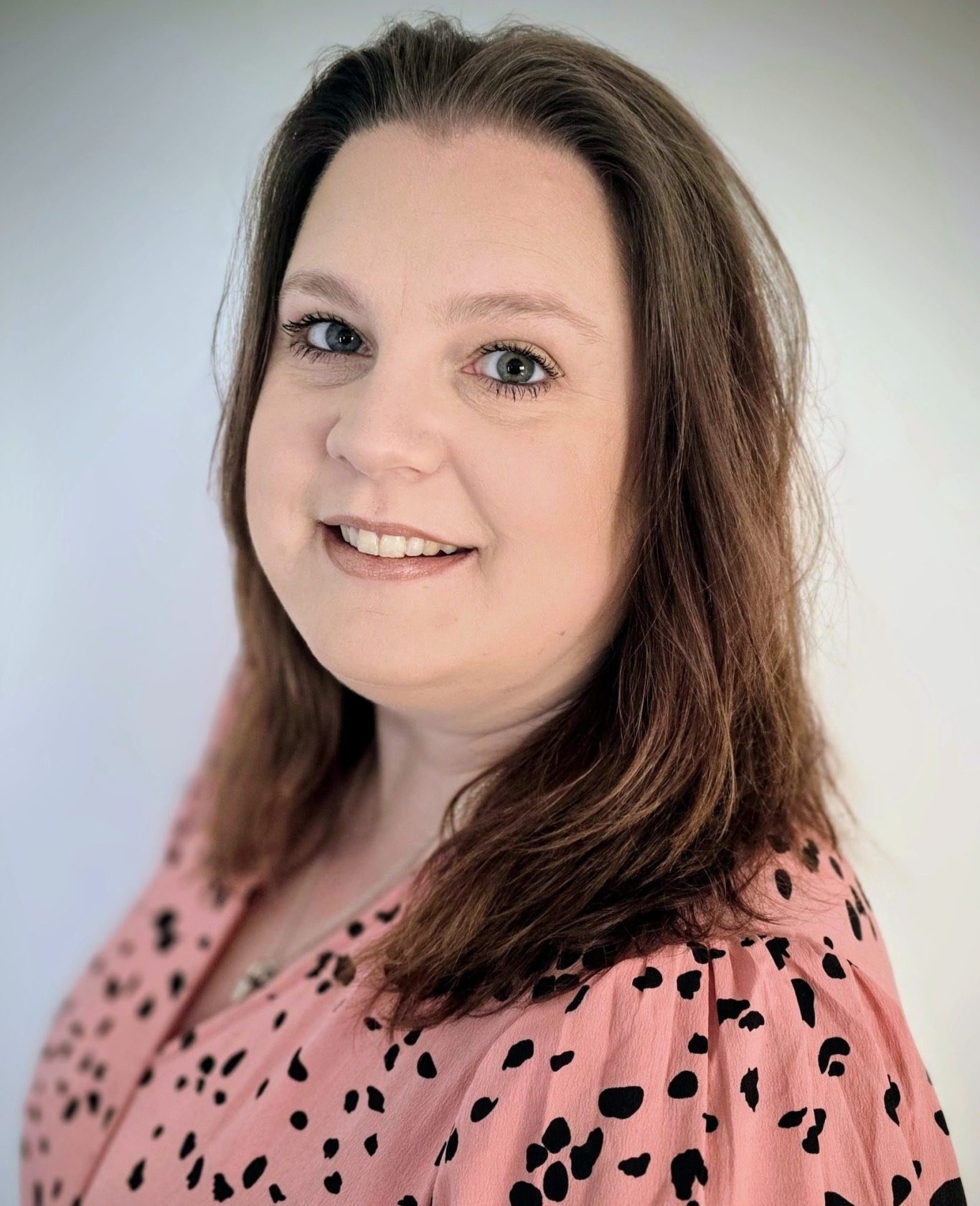 Smiling woman with long brown hair, wearing a pink shirt with black spots.
